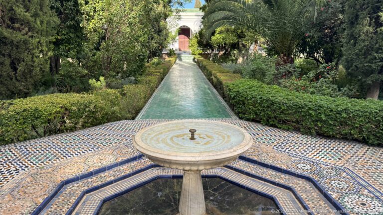 Pedestal fountain and tiled octagonal basin aligned with a long narrow water channel running between clipped garden hedges toward a doorway.