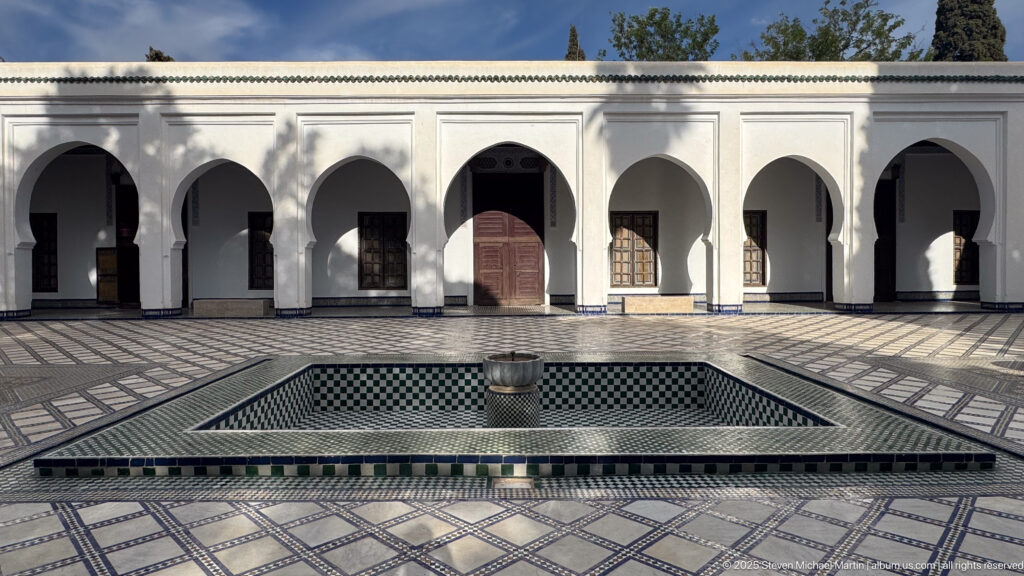 White arcaded courtyard with a central square tiled fountain basin, patterned pavement, and dark wooden doors and windows.