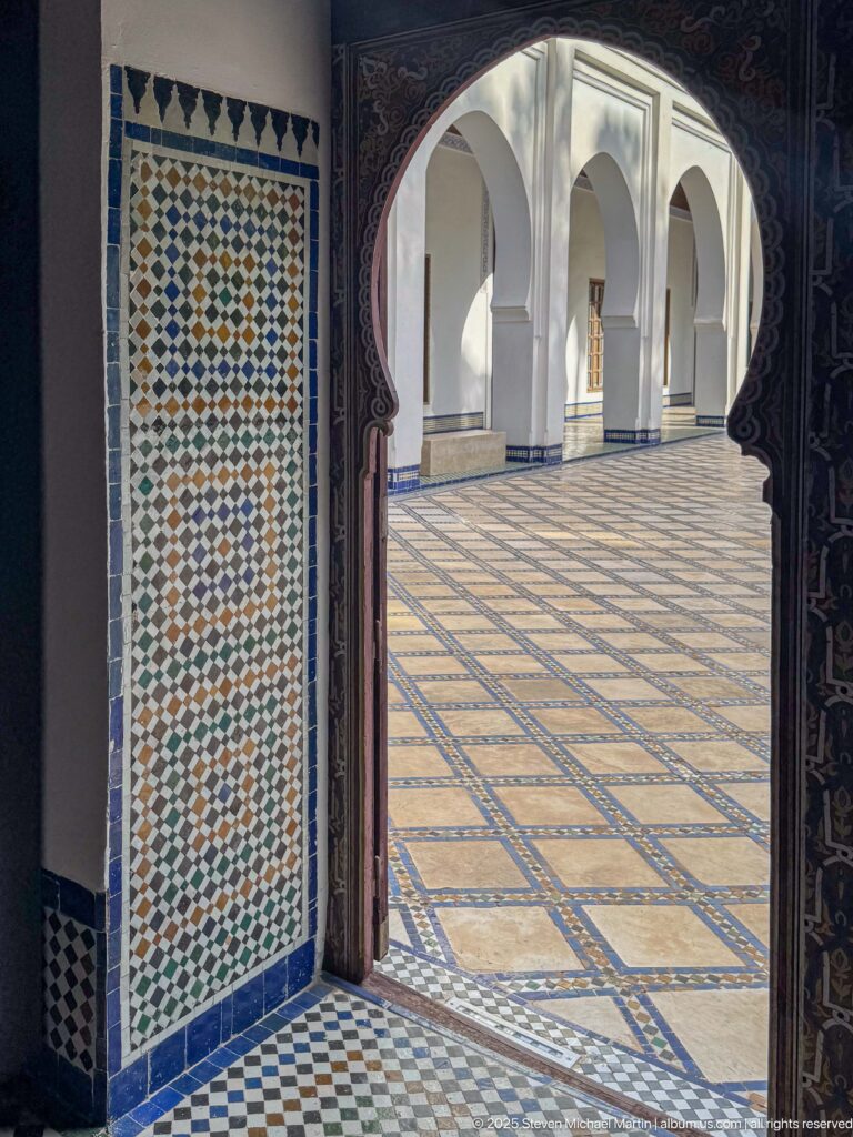 Dark carved doorway framing a bright arcaded courtyard with white arches, patterned paving, and multicolored geometric tilework at the threshold.
