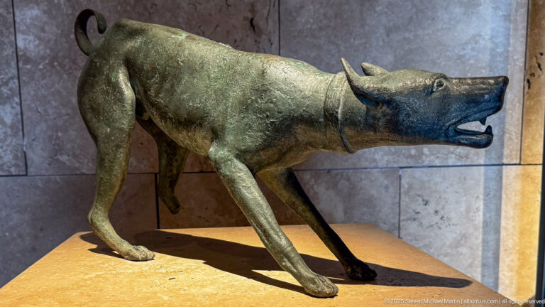 Patinated bronze quadruped statuette with pointed ears, long snout, open mouth, neck band, and slender legs.
