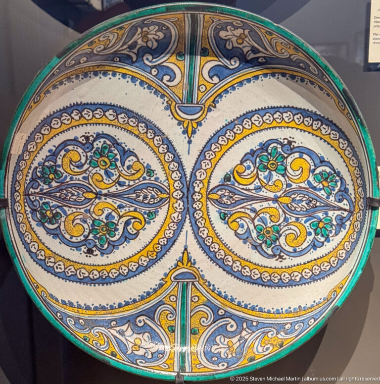 Large round ceramic platter with two symmetrical blue, yellow, green, and brown ornamental fields enclosed by a green rim.