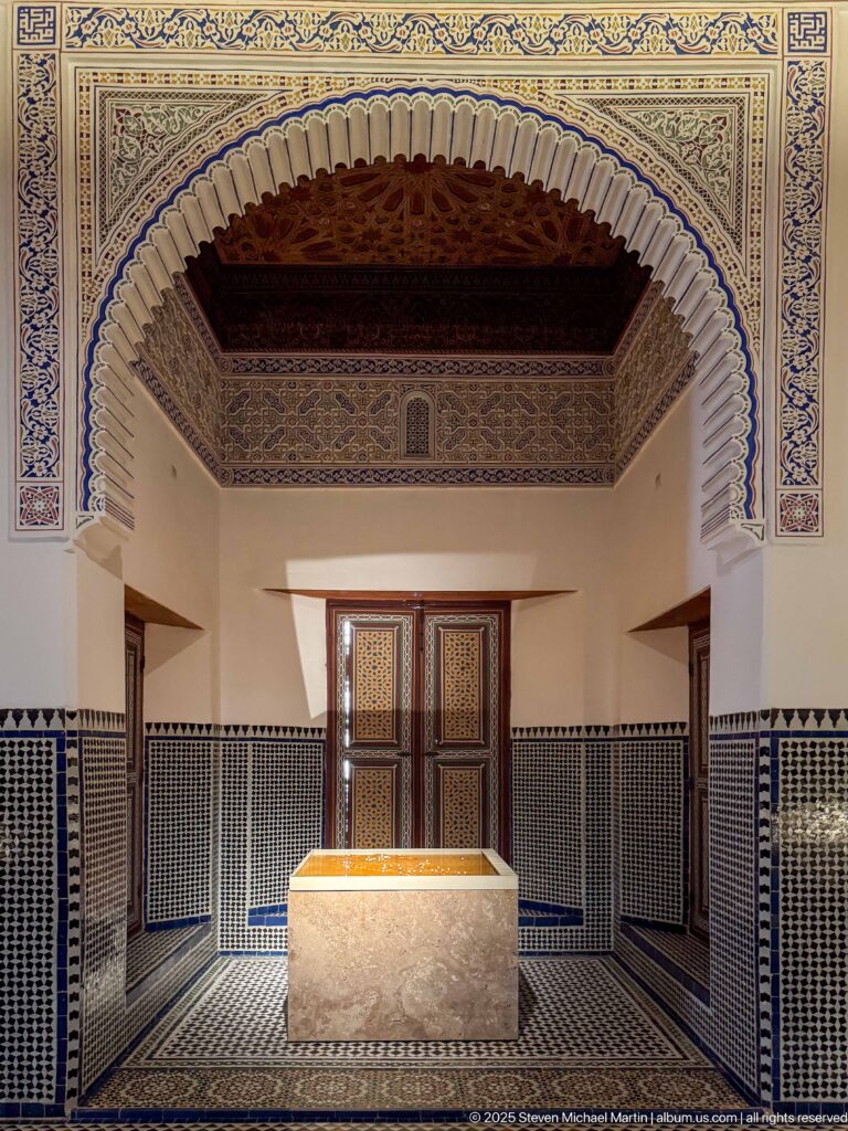 Interior room with scalloped arch, zellij wall panels, decorated upper walls, dark timber ceiling, wooden screen, and central pale plinth.