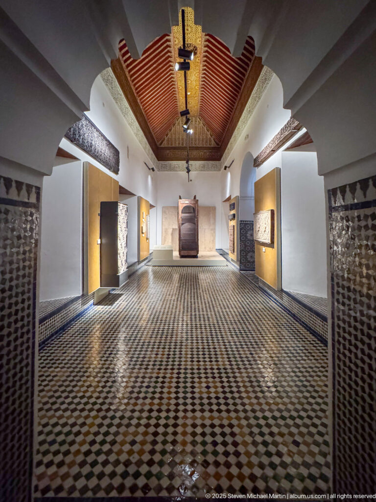 Long museum gallery with checker-pattern tile floor, wood ceiling, recessed displays, tiled wall panels, and a large object at the far end.