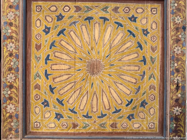Square polychrome panel with a large yellow-and-blue radiating rosette, geometric framing, and dark floral side borders.