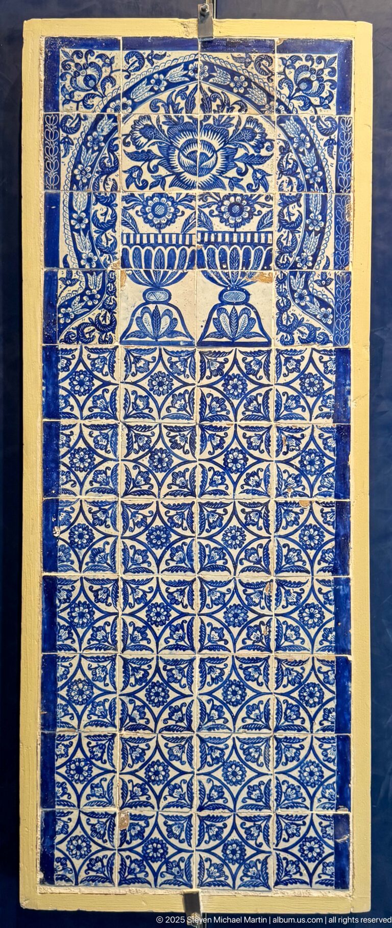 Rectangular blue-and-white ceramic tile panel with visible tile joints, dense vegetal patterns, and a symmetrical architectural motif across the top.