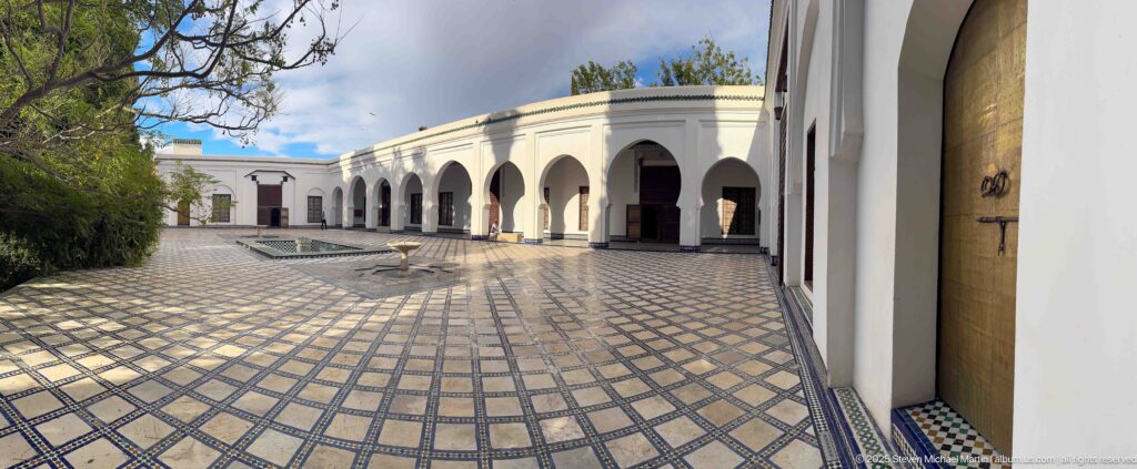 Wide museum courtyard with patterned paving, white arches, dark wooden doors, trees, and a small central fountain feature.
