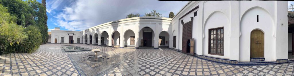 Small fountain in the foreground of a long patterned courtyard bordered by a white arcade, wooden doors, trees, and another basin beyond.