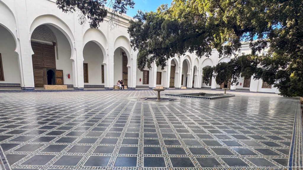 Large courtyard with black-and-white patterned paving, low central fountain, white arcades, dark wooden doors, and mature trees.
