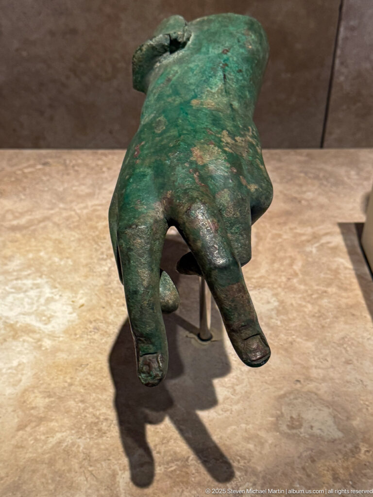 Small green-patinated bronze object shaped like a human hand with separated fingers and truncated wrist end.