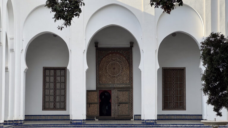 Large decorated wooden doorway centered beneath a white arch, flanked by two tall arched window bays and a narrow entrance below.
