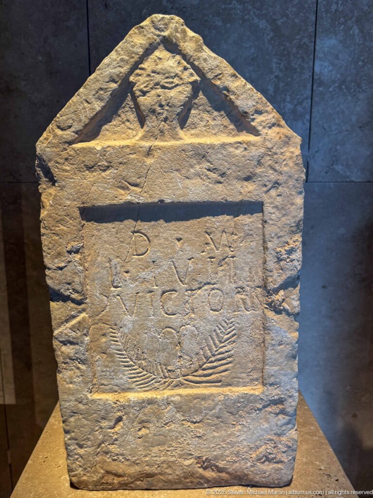 Weathered stone stele with triangular pediment, inset inscription panel, and central palm-like carved motif.