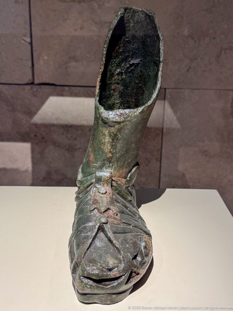 Patinated hollow bronze object shaped like a shoe or short boot with modeled toe and incised upper details.