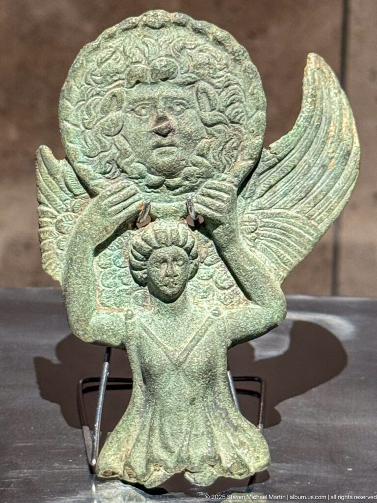 Green-patinated bronze appliqué with frontal human face, broad symmetrical wing-like forms, and pendant element below.