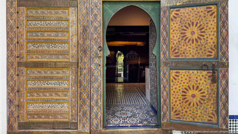 Polychrome wooden doorway with geometric panels and large rosettes opening toward a tiled interior gallery.