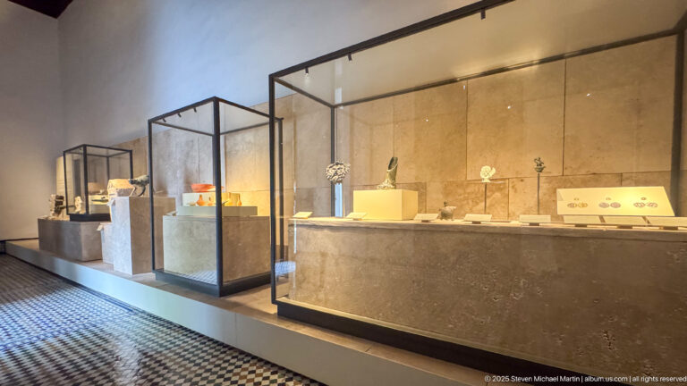 Long gallery with illuminated glass display cases holding small artifacts above a patterned tile floor.
