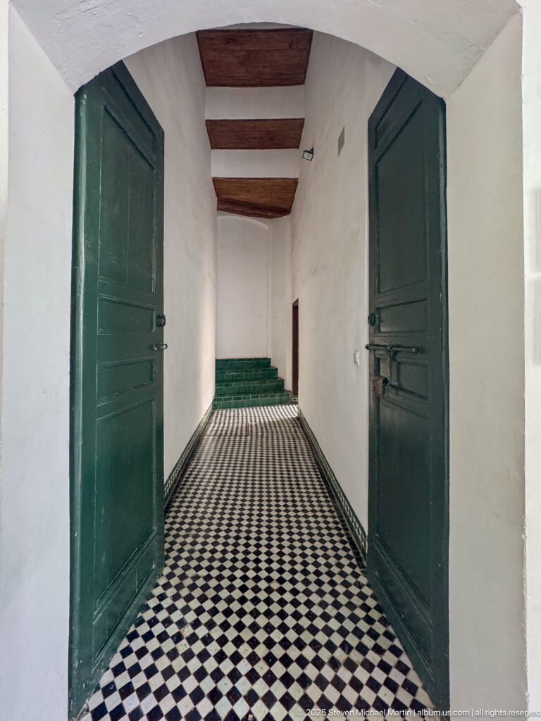 Narrow white corridor with green wooden doors, dark ceiling beams, black-and-white geometric floor tiles, and steps at the far end.