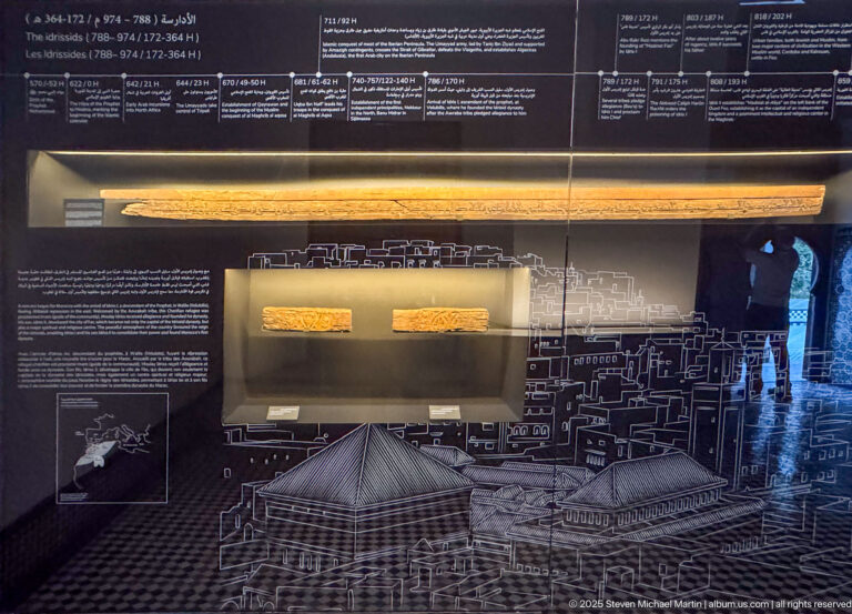 Dark museum display with long inscribed fragments, timeline text, white architectural diagrams, and a city-plan graphic.