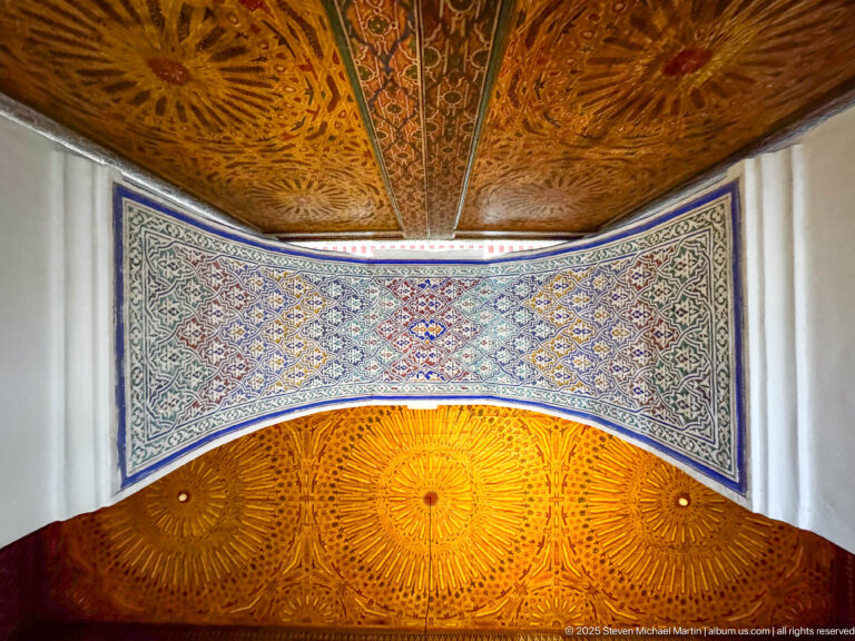 Decorated room with carved golden-brown wooden ceiling, large radial medallions, and blue geometric wall panel below.