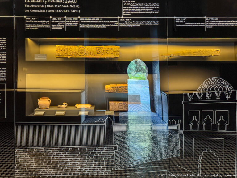Museum case with inscribed fragments, small ceramic vessels, carved pieces, and white line drawings of arches and buildings.