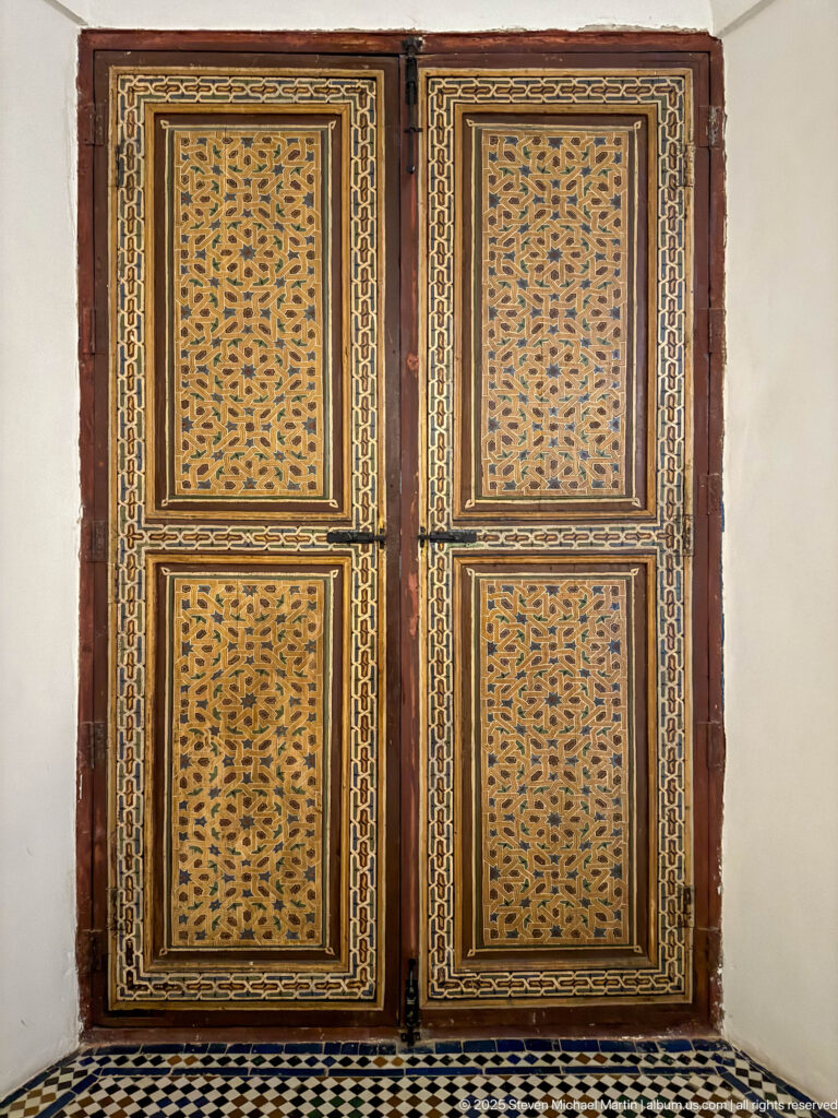 Pair of tall wooden doors with geometric openwork panels, painted borders, and a black-and-white tiled floor.