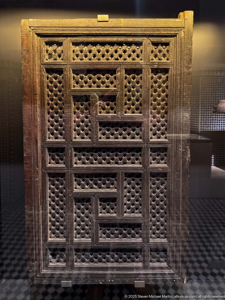 Rectangular wooden lattice panel with stepped framing and dense pierced geometric openings displayed upright in a museum case.
