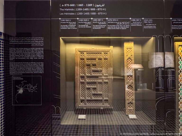 Museum case with a large wooden lattice panel and a narrow carved vertical wood element beneath a Marinid chronology.