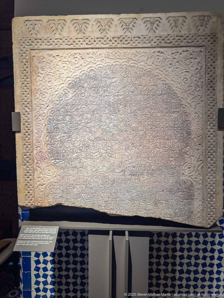 Large pale rectangular panel densely covered in Arabic script and framed by carved ornamental borders above blue-and-white tilework.