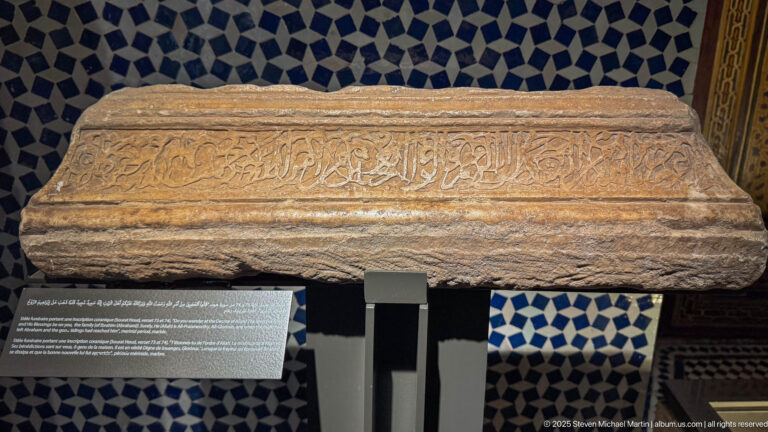 Tapered stone architectural block with a long carved Arabic inscription displayed against blue-and-white zellij.