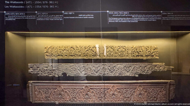 Museum case with several long carved architectural fragments bearing calligraphy, vegetal ornament, and geometric patterns beneath a Wattasid chronology.