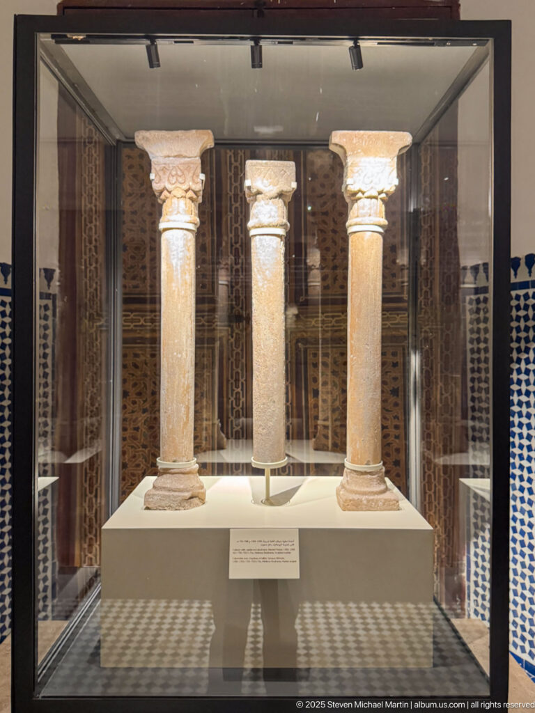 Three detached stone columns with carved capitals displayed upright together in a glass museum case.