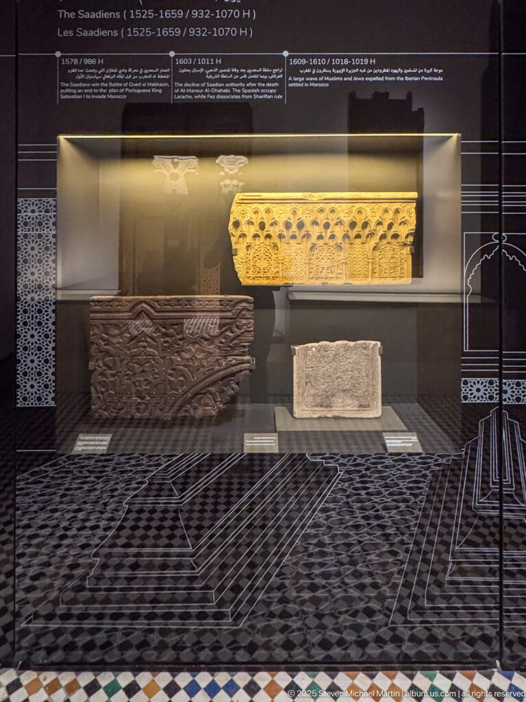 Three carved architectural fragments displayed at different heights in a glass case, including a long pale frieze and darker relief block.