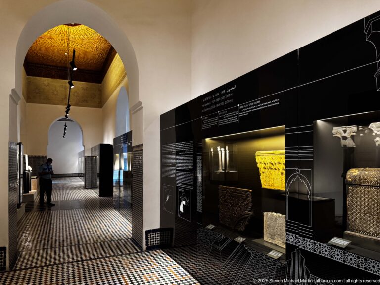 Arched museum corridor with patterned tile floor, dark exhibition cases, and a visitor standing midway down the gallery.