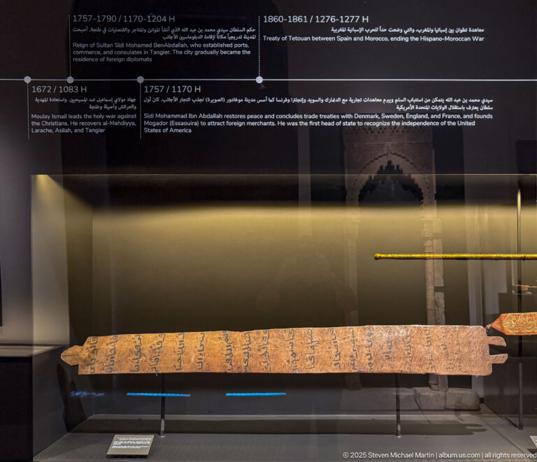 Long orange ceremonial textile with blue Arabic inscription displayed horizontally in a museum case.