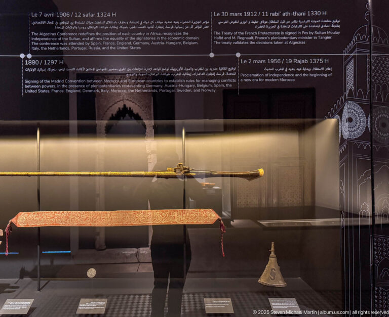 Museum case with a long ceremonial staff, orange inscribed textile, and small metal object beneath a diplomatic chronology.