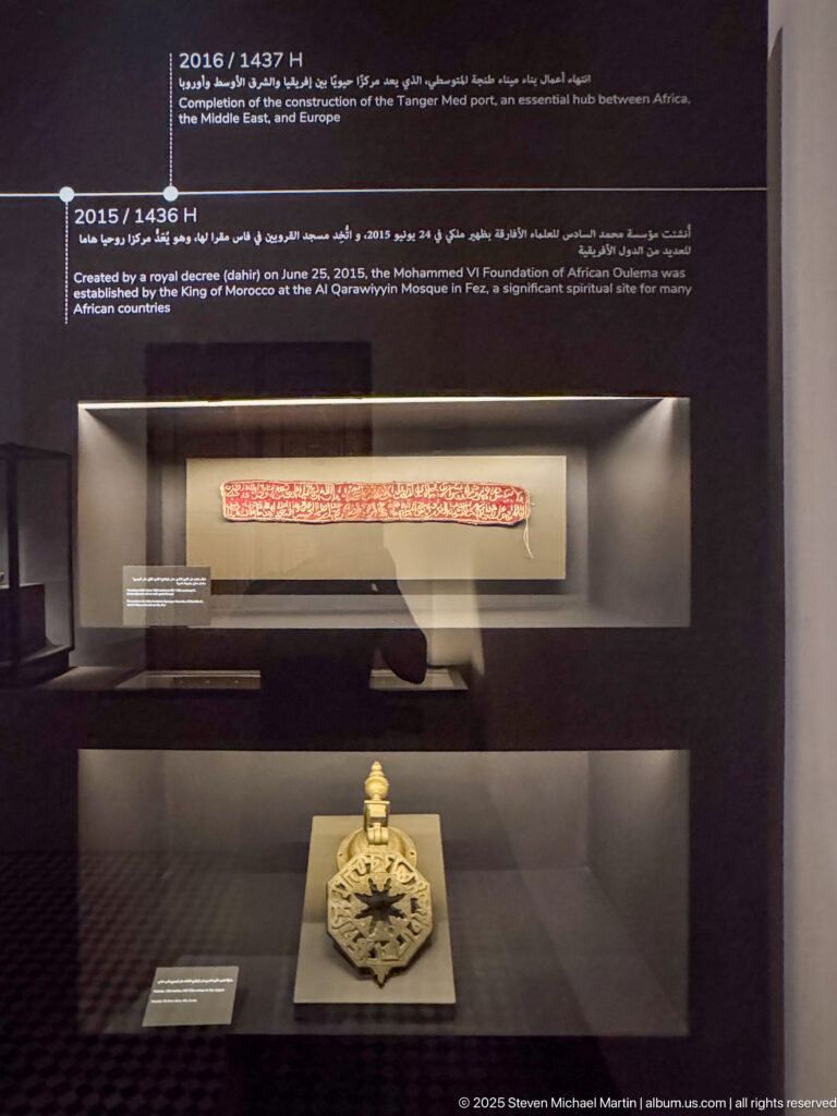 Short inscribed orange textile mounted above an ornate metal emblem or ceremonial fitting in a museum case.