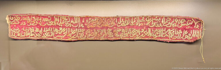 Long narrow red textile with several lines of dense gold Arabic calligraphy and frayed edges.
