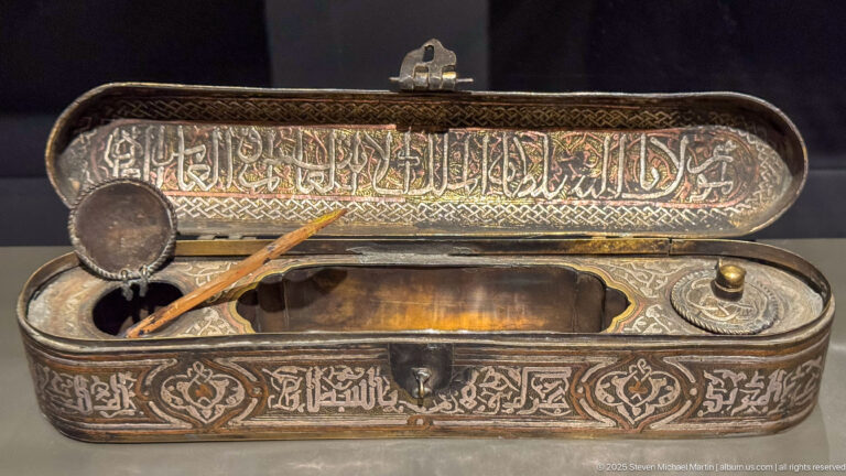 Open engraved metal qalamdan with Arabic inscriptions, reed pen, small inkwell, and fitted compartments for writing tools.