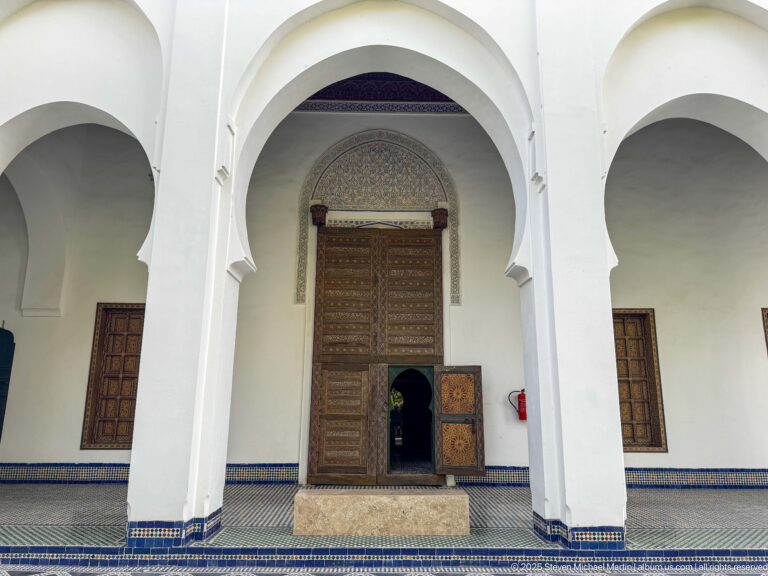 Courtyard arcade with white horseshoe arches, tall decorated wooden doorway, tiled floor, and low patterned wall band.