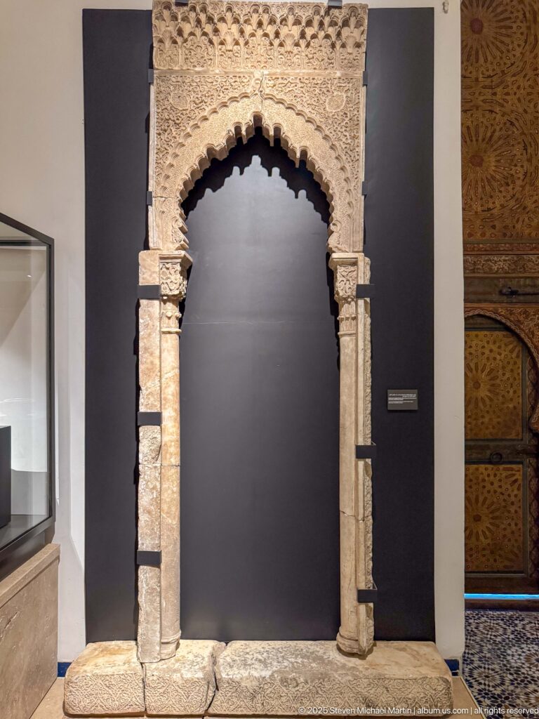 Freestanding pale carved arch with scalloped inner opening, slender columns, decorated capitals, and dense ornament across the crown.