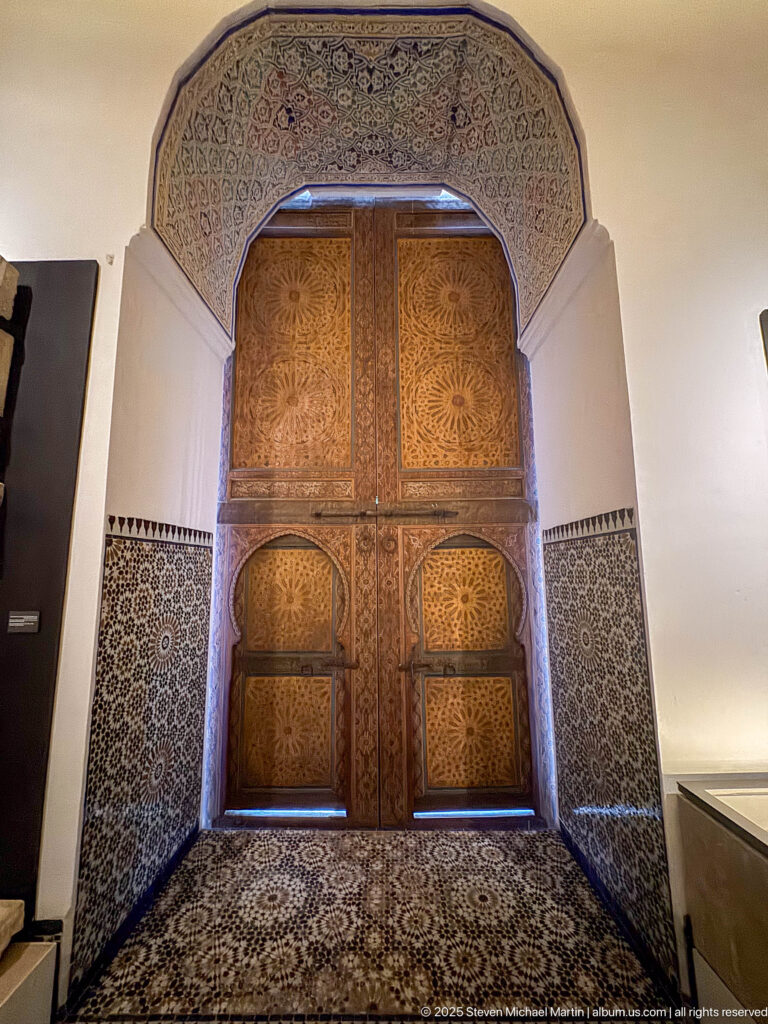 Double wooden doors in a horseshoe-arched opening with carved panels, decorated soffit, and blue-and-white zellij surround.