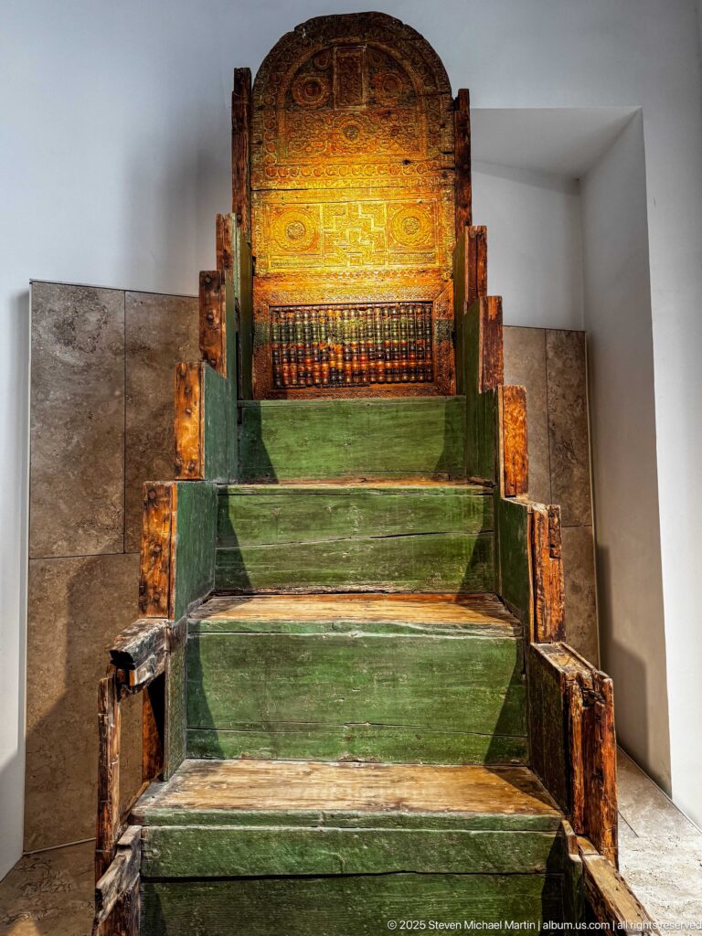 Large green-painted stepped wooden seat with a high rounded decorated back, projecting side elements, and worn surfaces.
