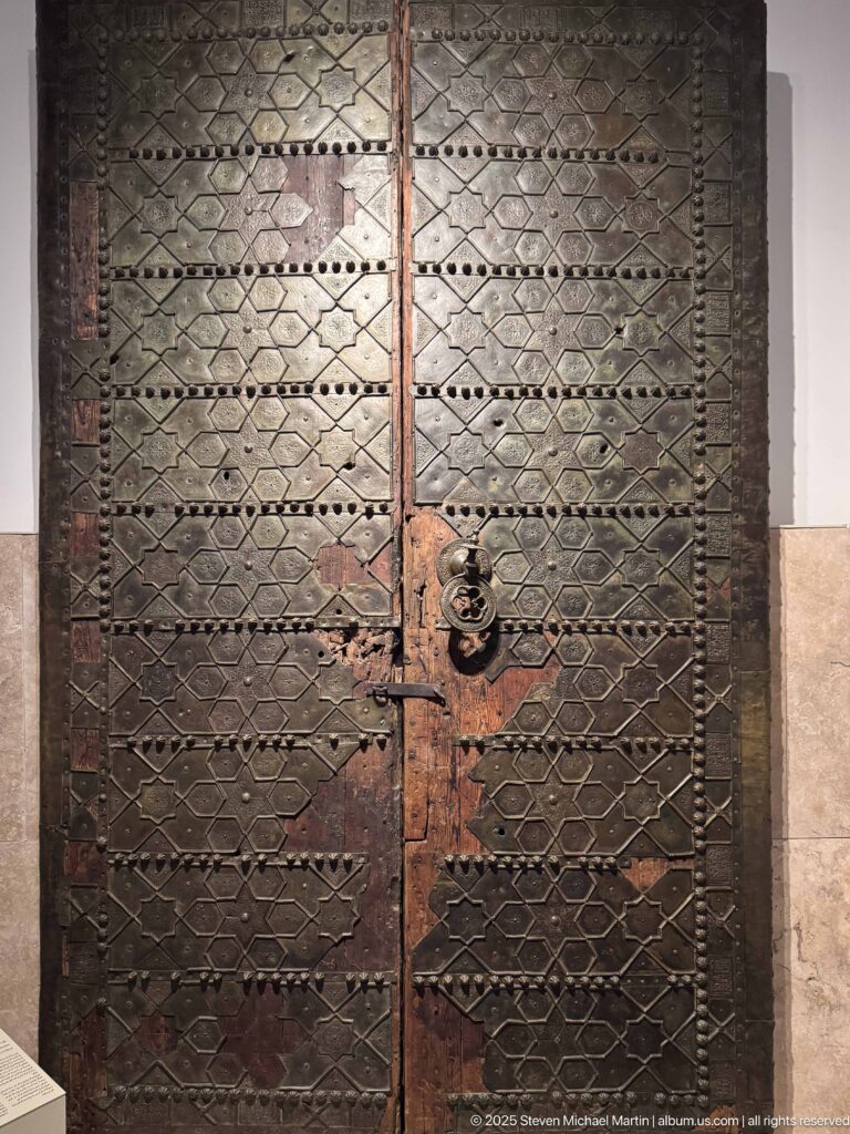 Dark double wooden doors with diamond-grid pattern, metal studs, worn central stile, and heavy ring handle.