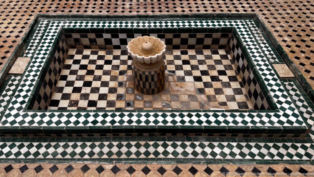 Square tiled fountain basin viewed from above, with checkerboard interior, geometric zellij border, and central scalloped fountain head.