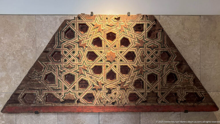 Trapezoidal carved wooden panel covered with interlaced star patterns and traces of red, green, gold, and dark paint.