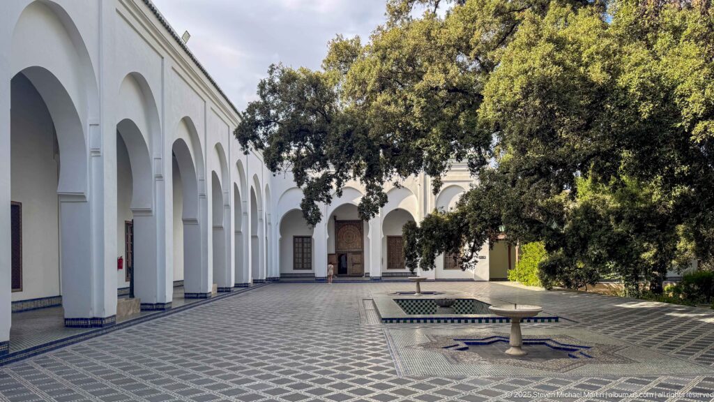 Wide museum courtyard with white arcades, mature tree, low fountain basin, patterned paving, and tiled wall borders.