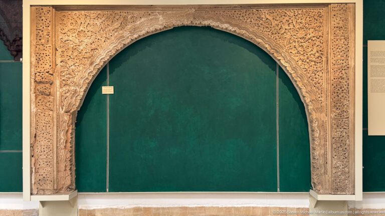 Pale carved horseshoe arch with dense vegetal and geometric ornament displayed against a dark green museum wall.