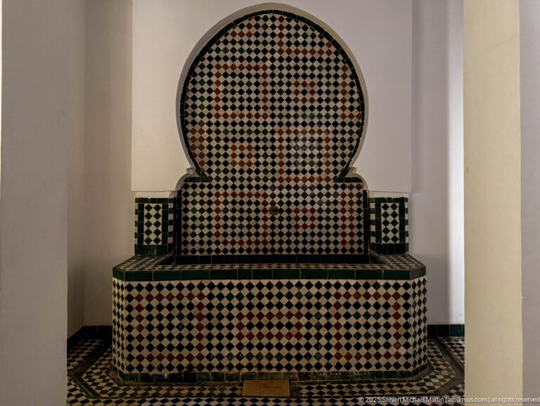 Arched wall basin faced with black, white, green, and reddish checkerboard zellij above a rectangular tiled trough.