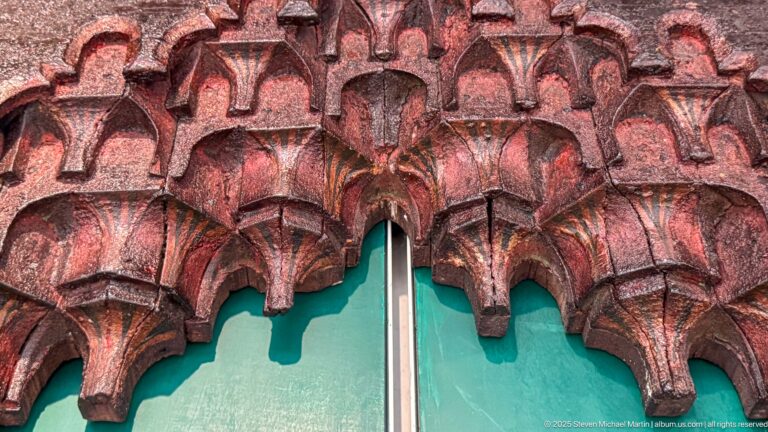 Close-up of red-painted wooden muqarnas with repeated pointed niche-like cells, faceted projections, and worn pigment.