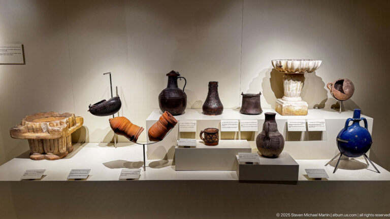 Museum case with dark glazed jugs, small cups, orange vessels, a cobalt-blue pitcher, and a pale fluted ceramic fragment.