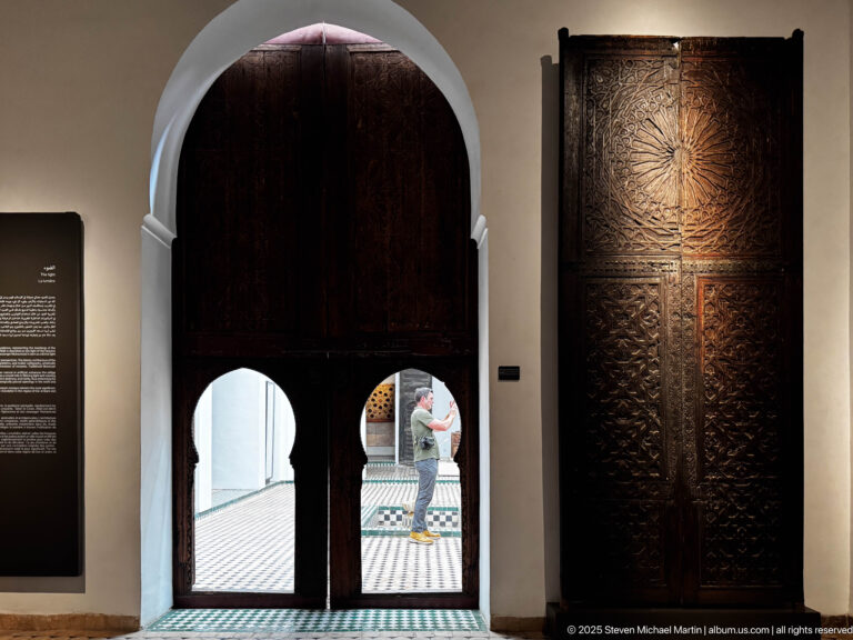 Dark carved wooden doors with two lower arched openings, adjacent carved panel, tiled floor, and a visitor beyond.